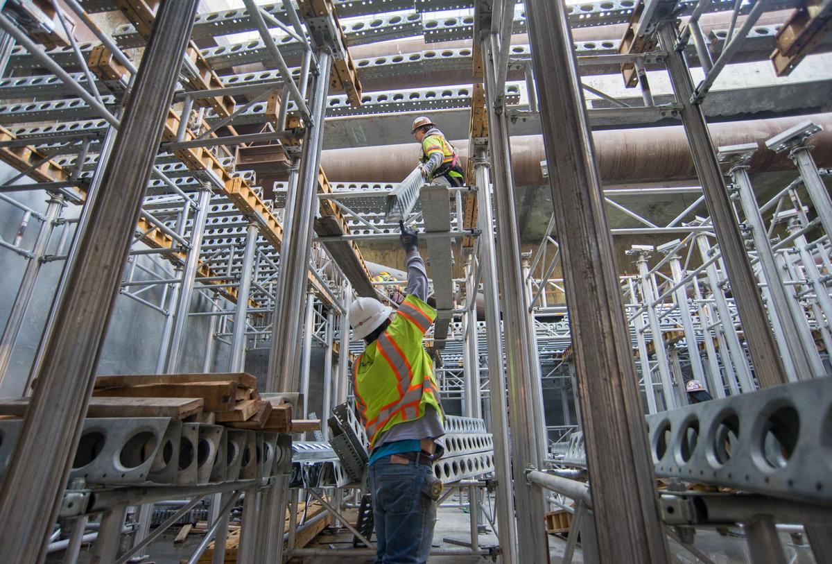 Workers assemble steel scaffolding and concrete forms inside an industrial construction site.