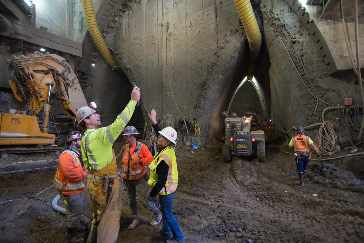 Workers in safety gear conduct air quality testing inside underground tunnel construction site with large boring equipment visible.