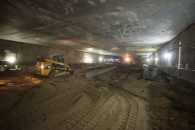 Underground station platform under construction with Bobcat equipment and excavated material during utility relocation work.