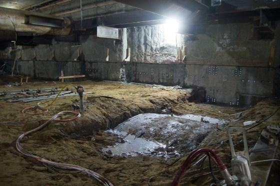 Underground station box construction site with excavated earth, water pooling, and red hoses beneath concrete ceiling and industrial lighting.