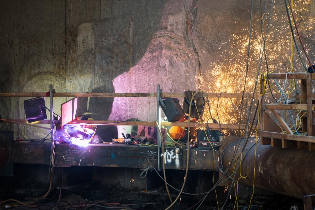 Worker welding underground infrastructure with bright sparks and purple light illuminating the industrial construction site.