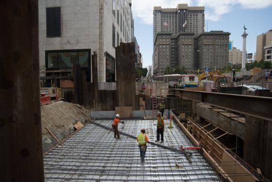 Workers installing rebar lattice on underground station roof deck surrounded by construction barriers and city buildings.