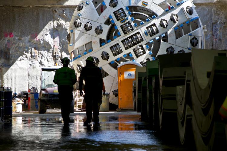Two workers stand in a wet tunnel beneath a large tunnel boring machine under assembly, with blue and white mechanical components visible overhead.