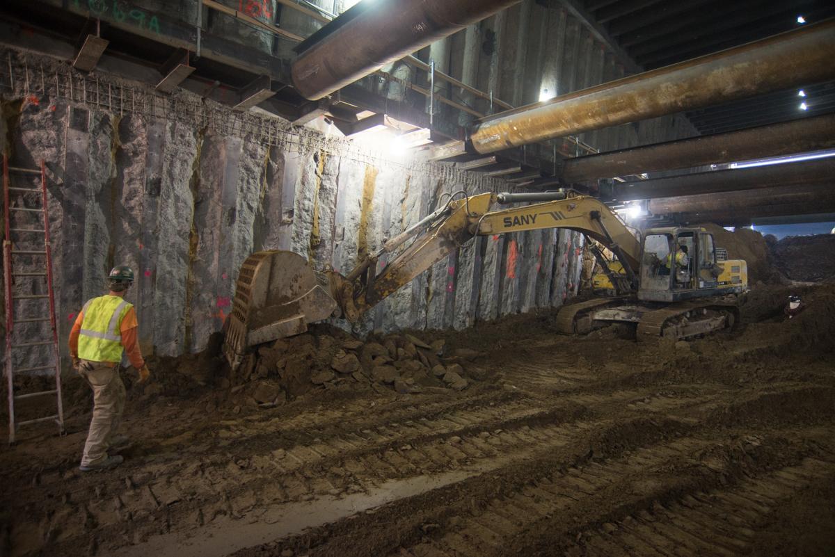 Excavator collecting concrete debris inside underground station box with worker in safety vest nearby.