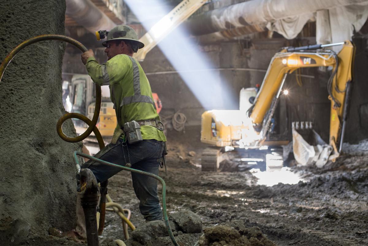 Worker operating high-pressure water hose in underground transit tunnel excavation with yellow machinery visible.