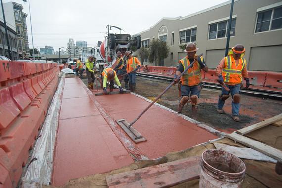 Construction workers in safety vests laying cement between transit tracks at an urban work site.