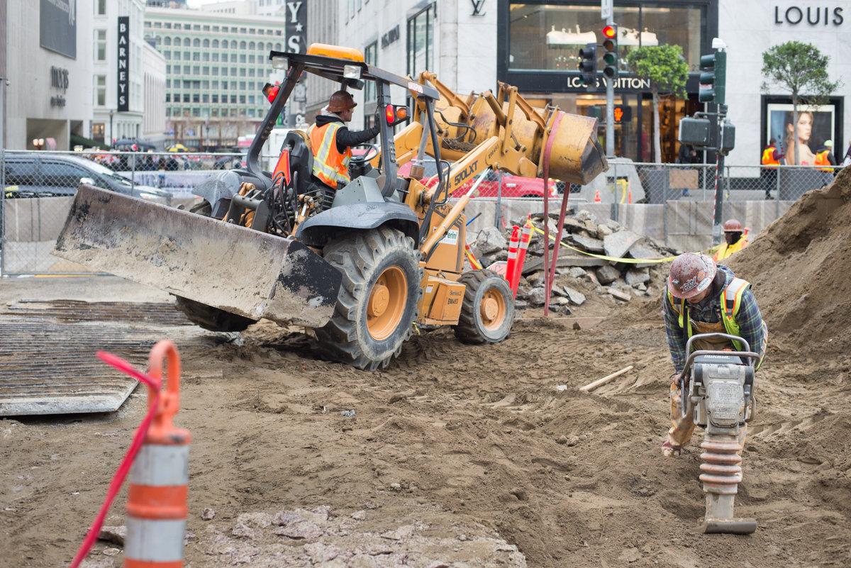 Construction crew operating heavy equipment for soil compaction and utility work on urban street.