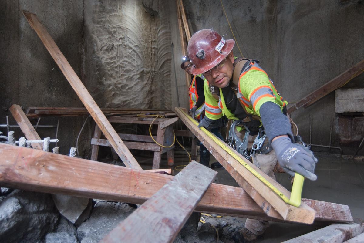 Worker in safety gear measuring wooden beam inside concrete structure during construction.
