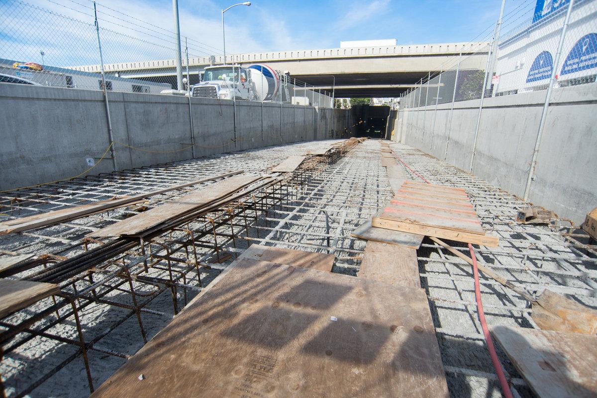 Construction site showing reinforced steel rebar and concrete foundation work inside a tunnel portal beneath an overpass.