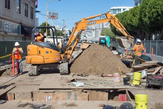 Construction crew operates excavator at Central Subway project site on city street.