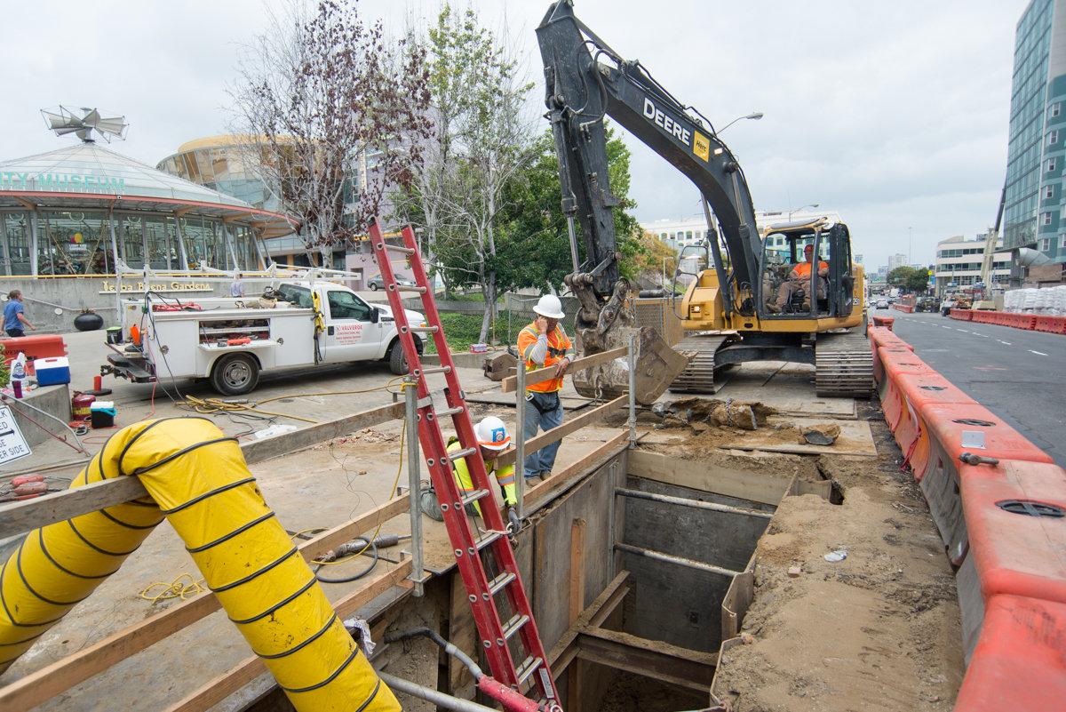 Construction site at 4th and Howard intersection with excavator, utility tunnel, and workers in safety gear.