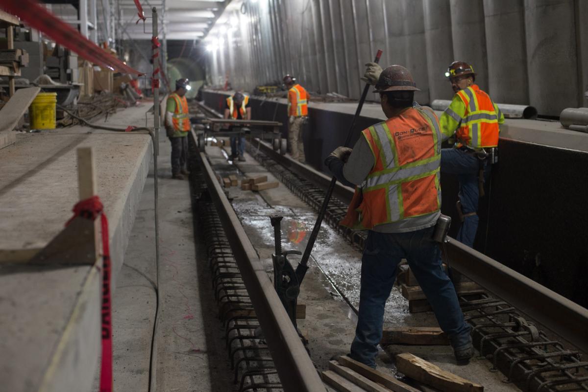 Construction workers in safety vests install utility conduits and lighting in a transit tunnel.