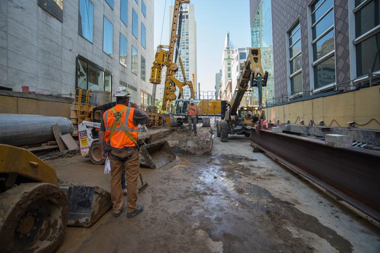 Construction crew in orange safety vest working on underground utility replacement in downtown street corridor between tall buildings.