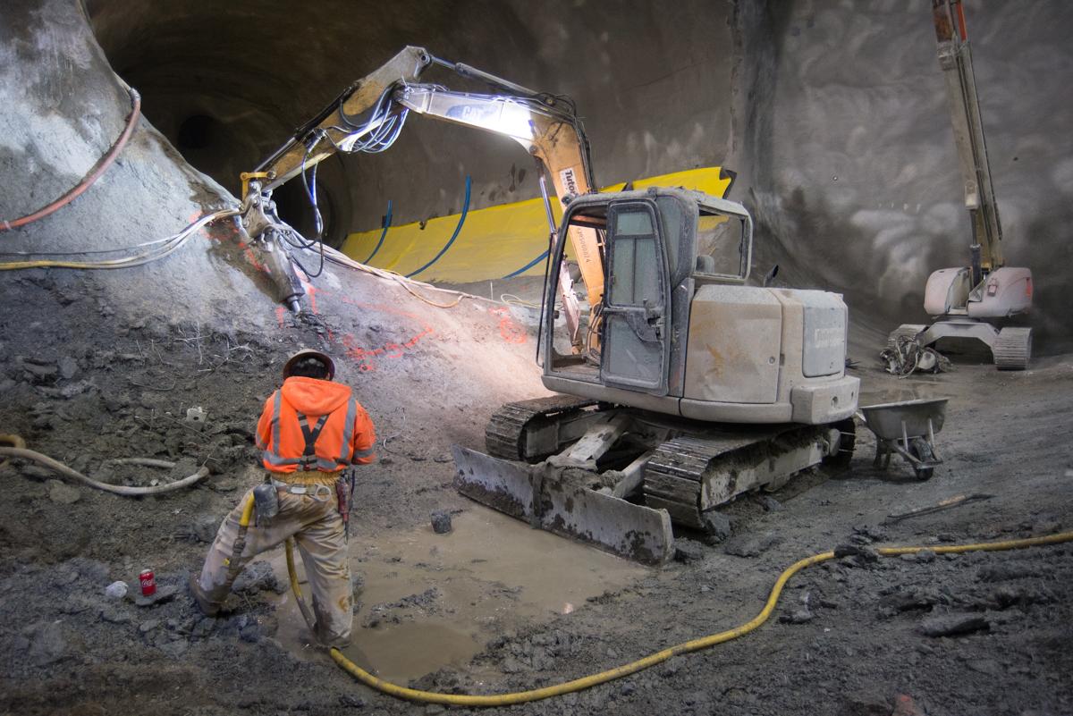 Excavator with jackhammer attachment removes shotcrete from tunnel wall while worker in safety vest controls dust spray.