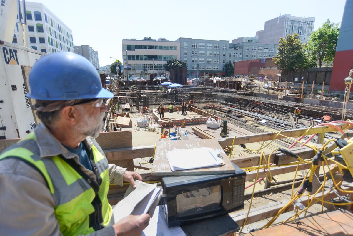 Construction worker in hard hat and safety vest reviews plans at active transit station construction site.