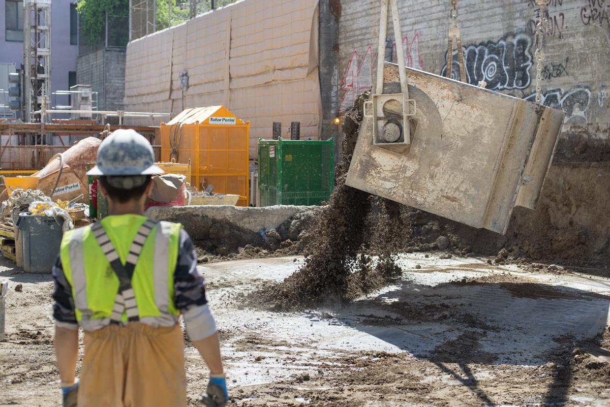 Worker in safety gear observing concrete bucket dumping excavated material at underground station construction site.