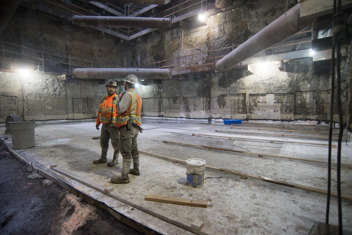 Two workers in safety gear inspect a concrete platform cavern with exposed pipes and construction materials during transit station repairs.