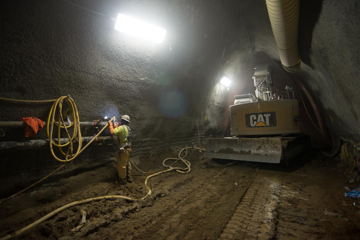 Worker manages air hoses while CAT excavator with grinding wheel operates in underground platform cavern.