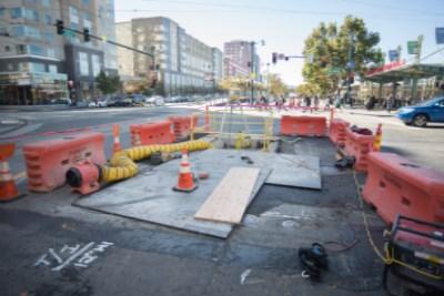 Urban street construction site with red barricades, traffic cones, and equipment blocking roadway near buildings.