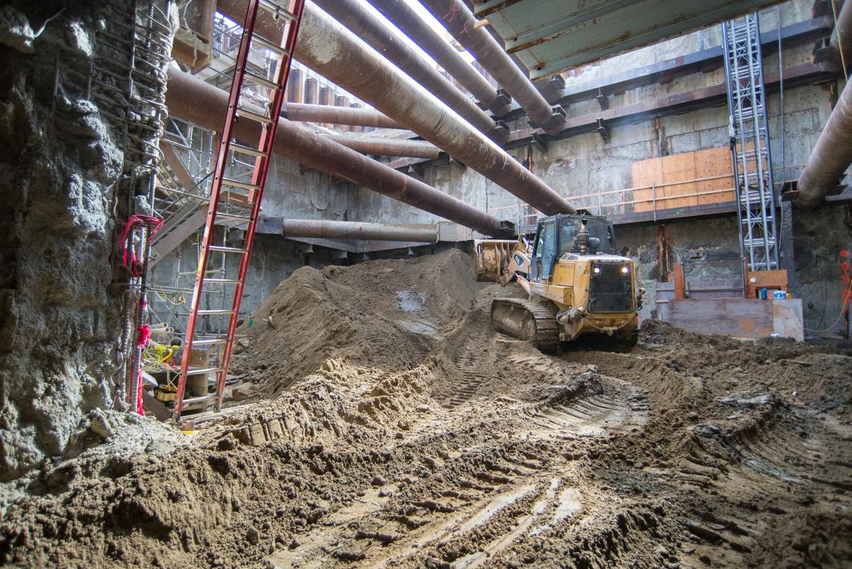 Construction equipment and excavated soil inside underground station box with concrete structural elements and red safety ladder visible.