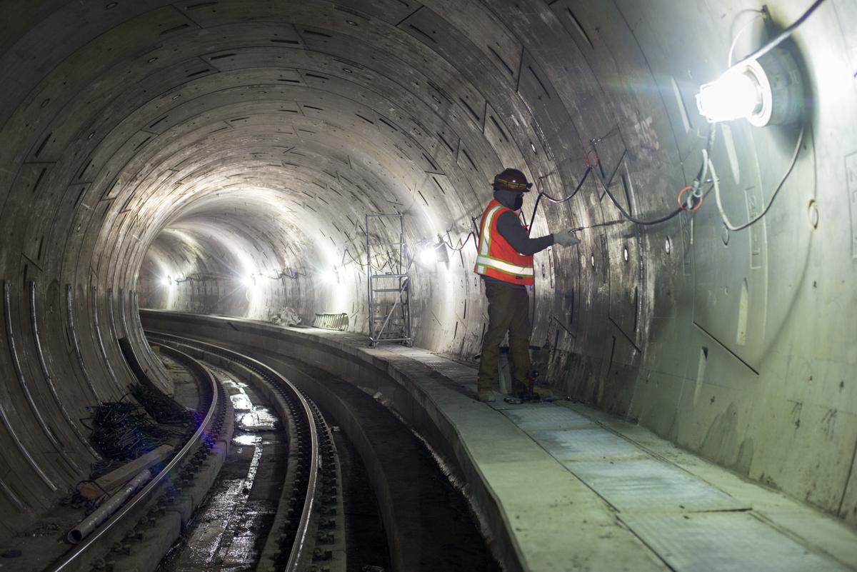 Worker in safety vest inspecting tunnel wall inside curved section of subway tunnel with railroad tracks visible.