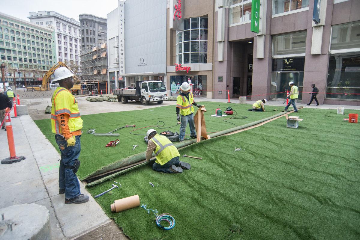 Construction workers in safety vests install artificial turf on a downtown San Francisco street.