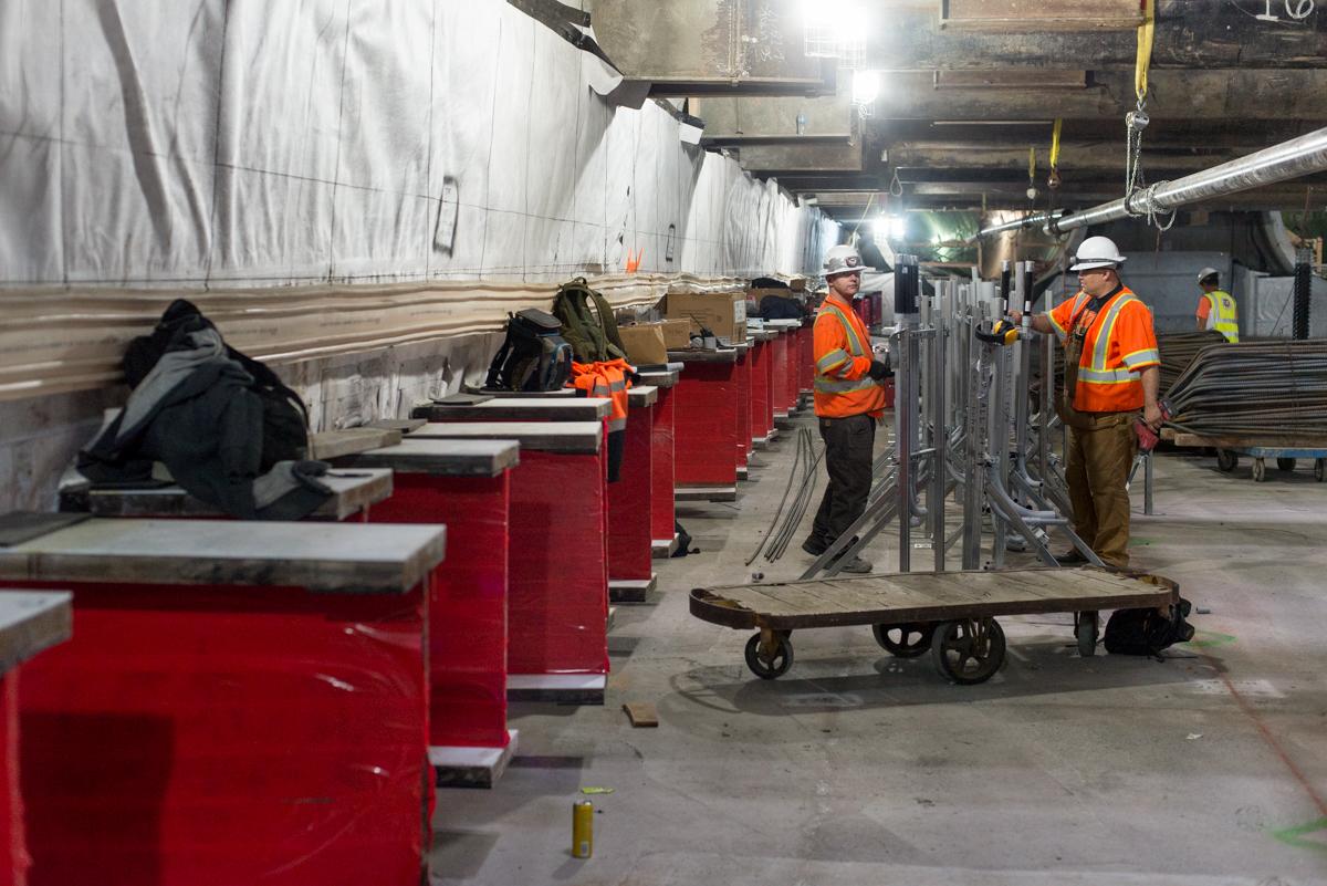 Workers in safety vests install utility conduits and pipes in an underground transit station construction area.