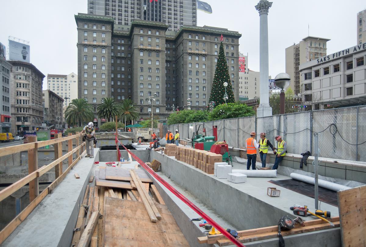 Construction site at Union Square with excavation pit, wooden bracing, workers in safety gear, and San Francisco buildings in background.