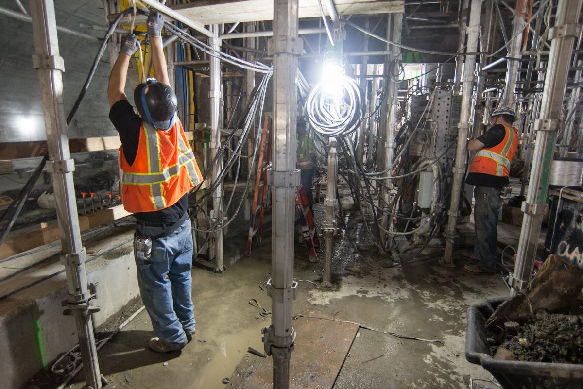Workers in safety vests install electrical cables and lighting equipment inside a transit station under construction.