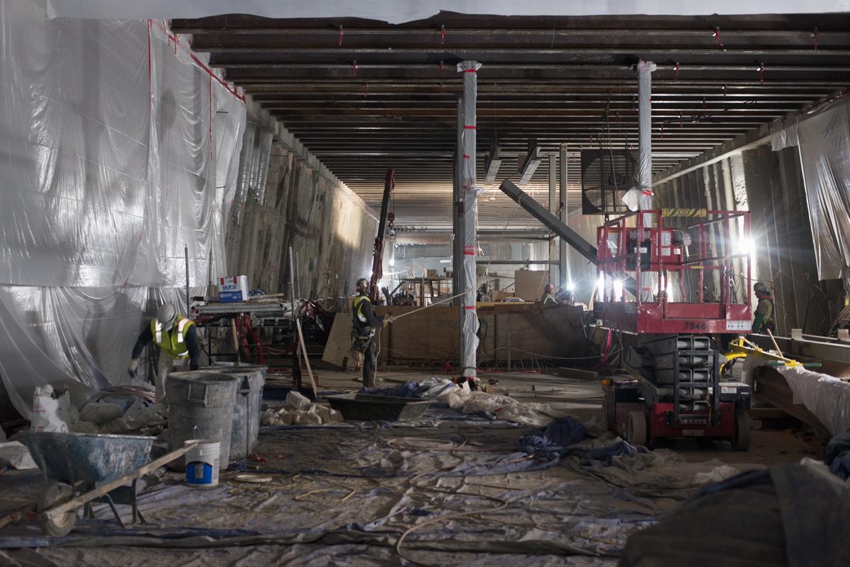 Construction workers install steel bracing beams inside a transit station mezzanine level under plastic sheeting.