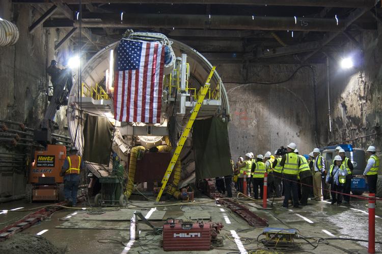 Workers in hard hats and safety vests observe a tunnel boring machine decorated with an American flag in an underground tunnel.