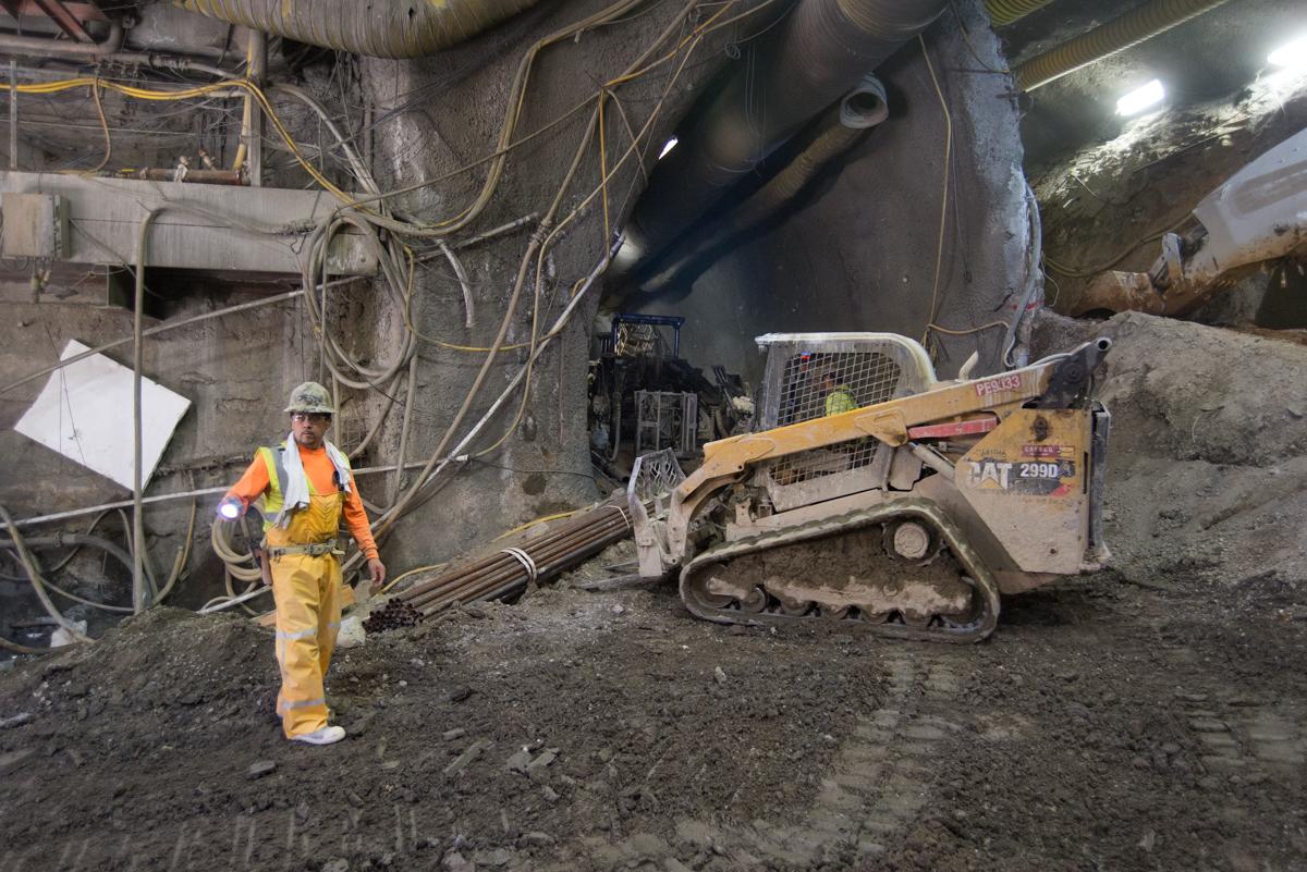 Worker in safety gear directing a tracked excavator inside an underground station cavern during construction.