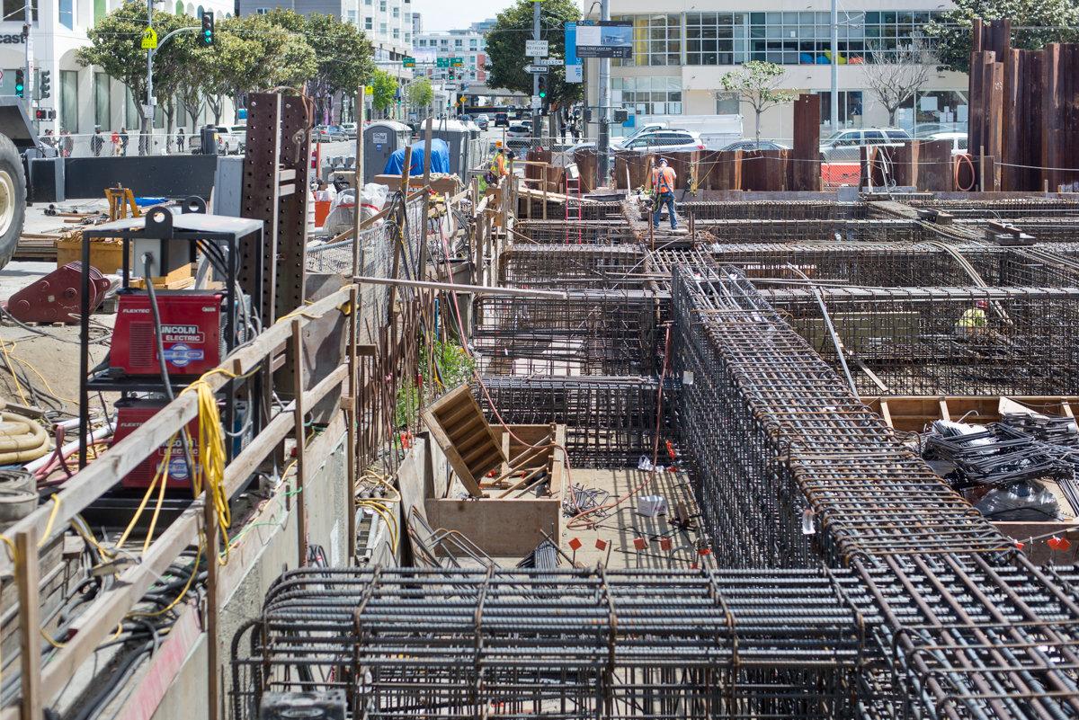 Rebar cages arranged in rows at a construction site, viewed from street level with city buildings in background.