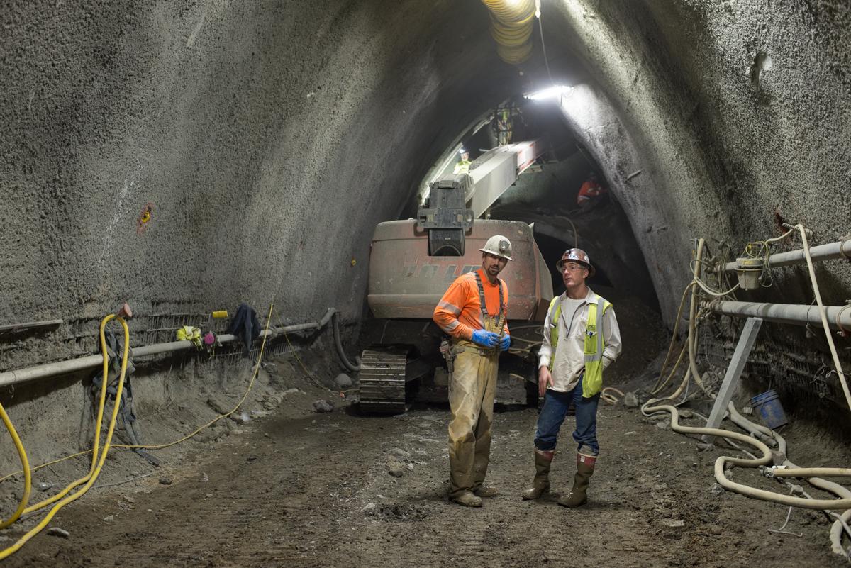 Two workers in hard hats and safety vests stand in an underground tunnel with a bulldozer and construction equipment during platform cavern excavation.
