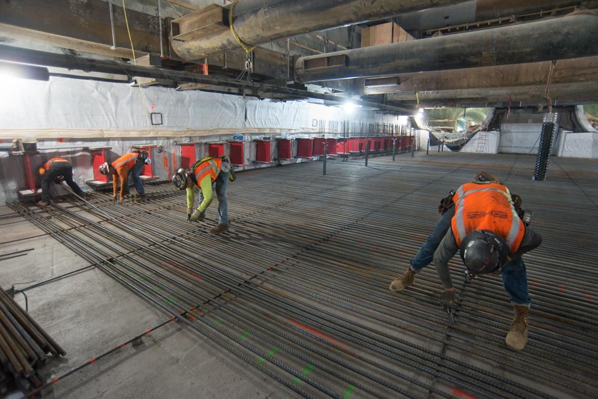 Workers in safety vests install rebar reinforcement on a concrete floor beneath an elevated structure.