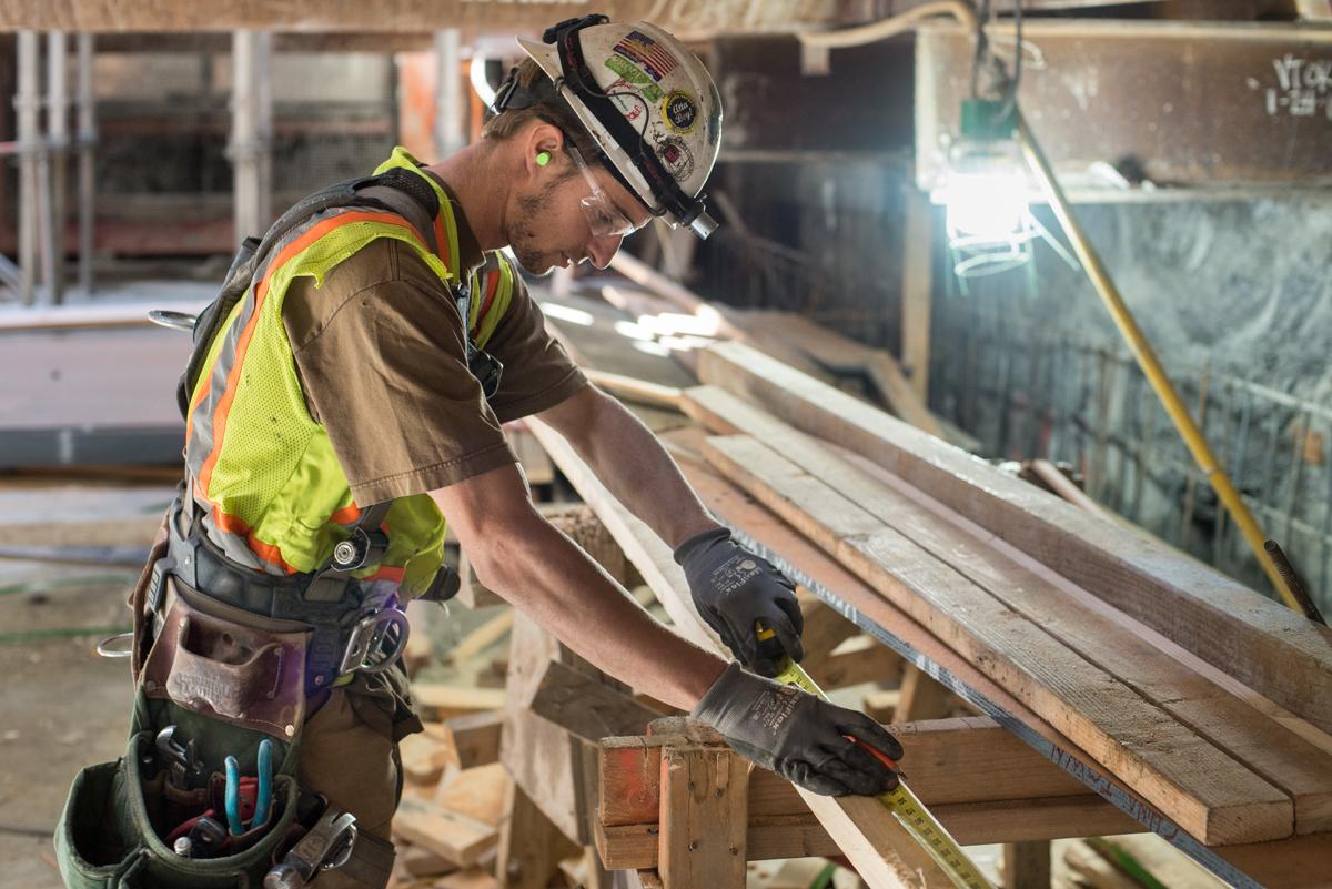 Construction worker in safety gear measuring wooden framing for concrete forms at transit station.