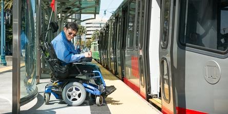Person in blue jacket on mobility device boarding accessible area of transit vehicle at platform.