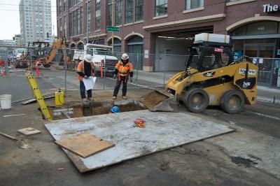 Construction crew performs underground utility work at street site with skid steer loader and excavation equipment near urban buildings.