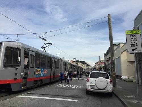 NJ Transit light rail train stopped at street crossing with pedestrians and vehicles nearby.