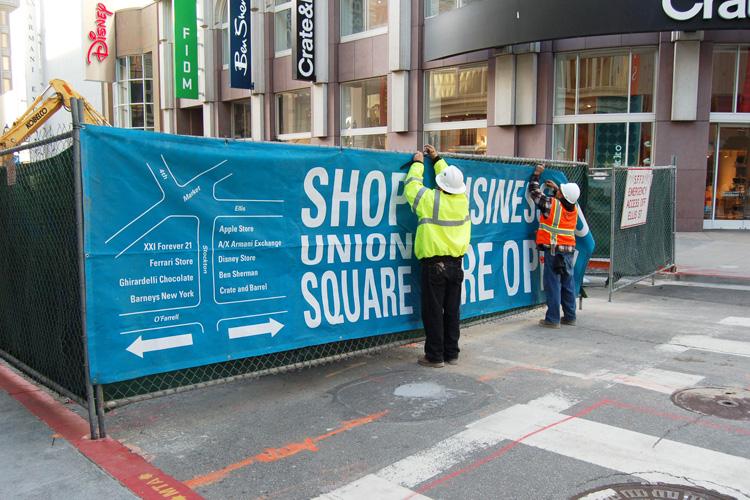 Construction site banner on Stockton Street with directional map and store listings, workers in safety vests nearby.