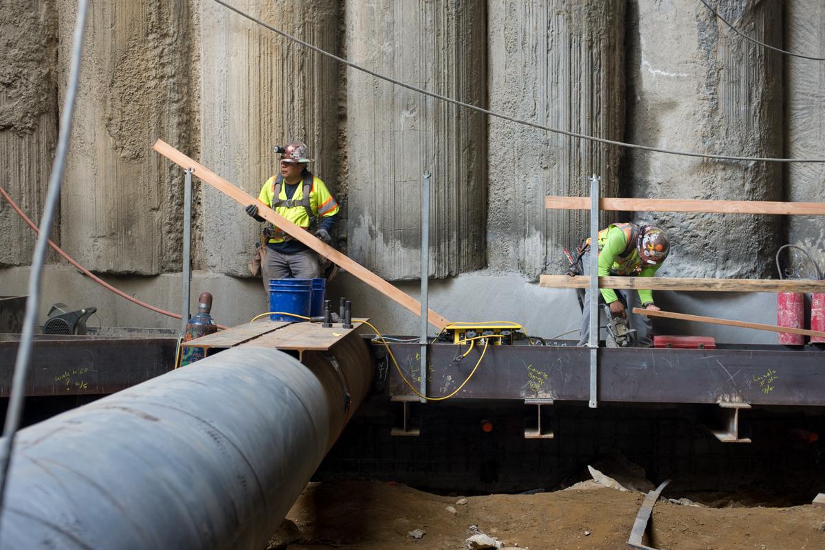 Workers in safety vests install temporary bracing and railings inside an underground transit station excavation site.