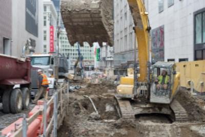 Excavation equipment and dump truck unloading soil on Stockton Street construction site surrounded by urban buildings.