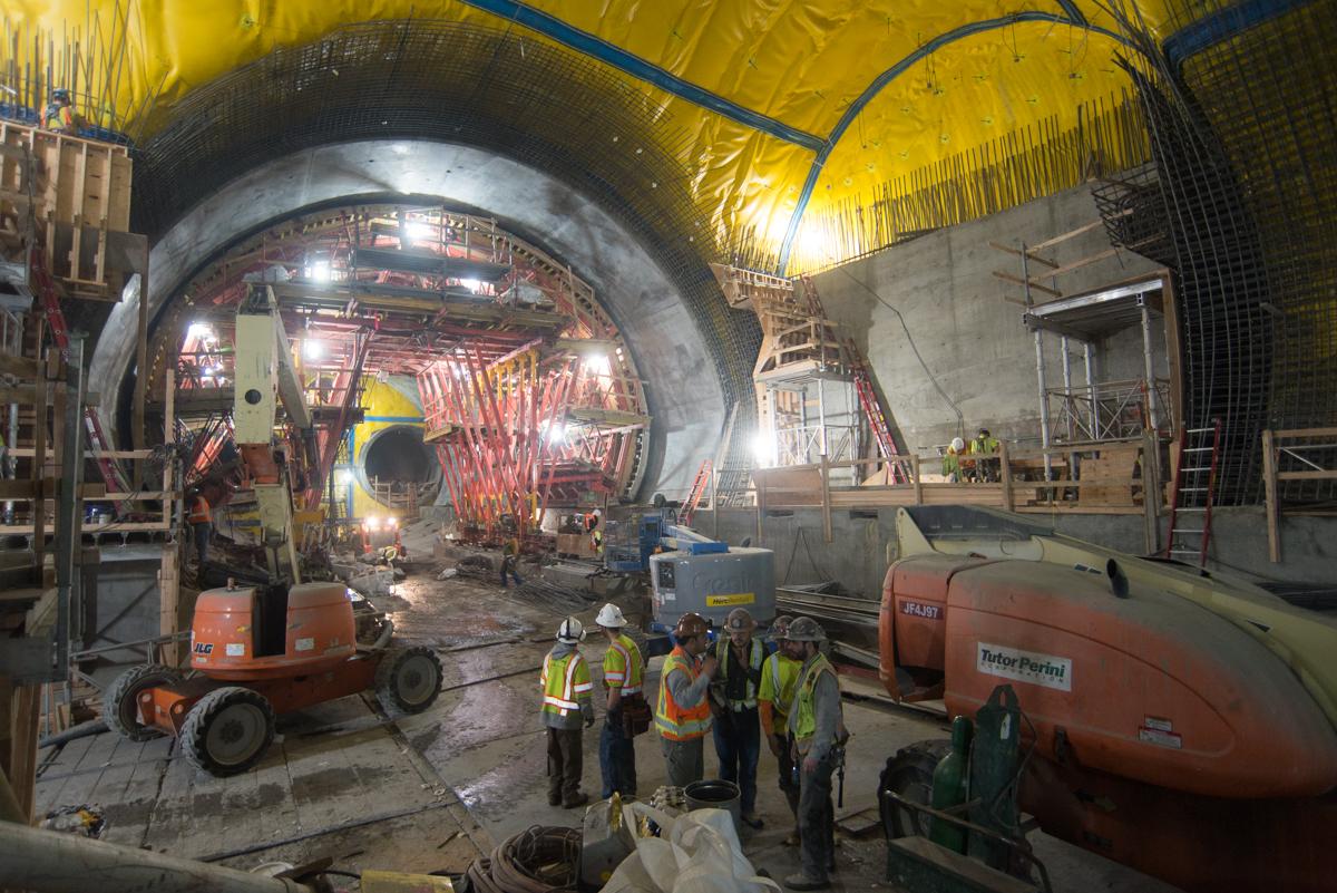 Construction crew in yellow safety vests gathered inside large underground tunnel cavern with heavy equipment and yellow protective sheeting overhead.