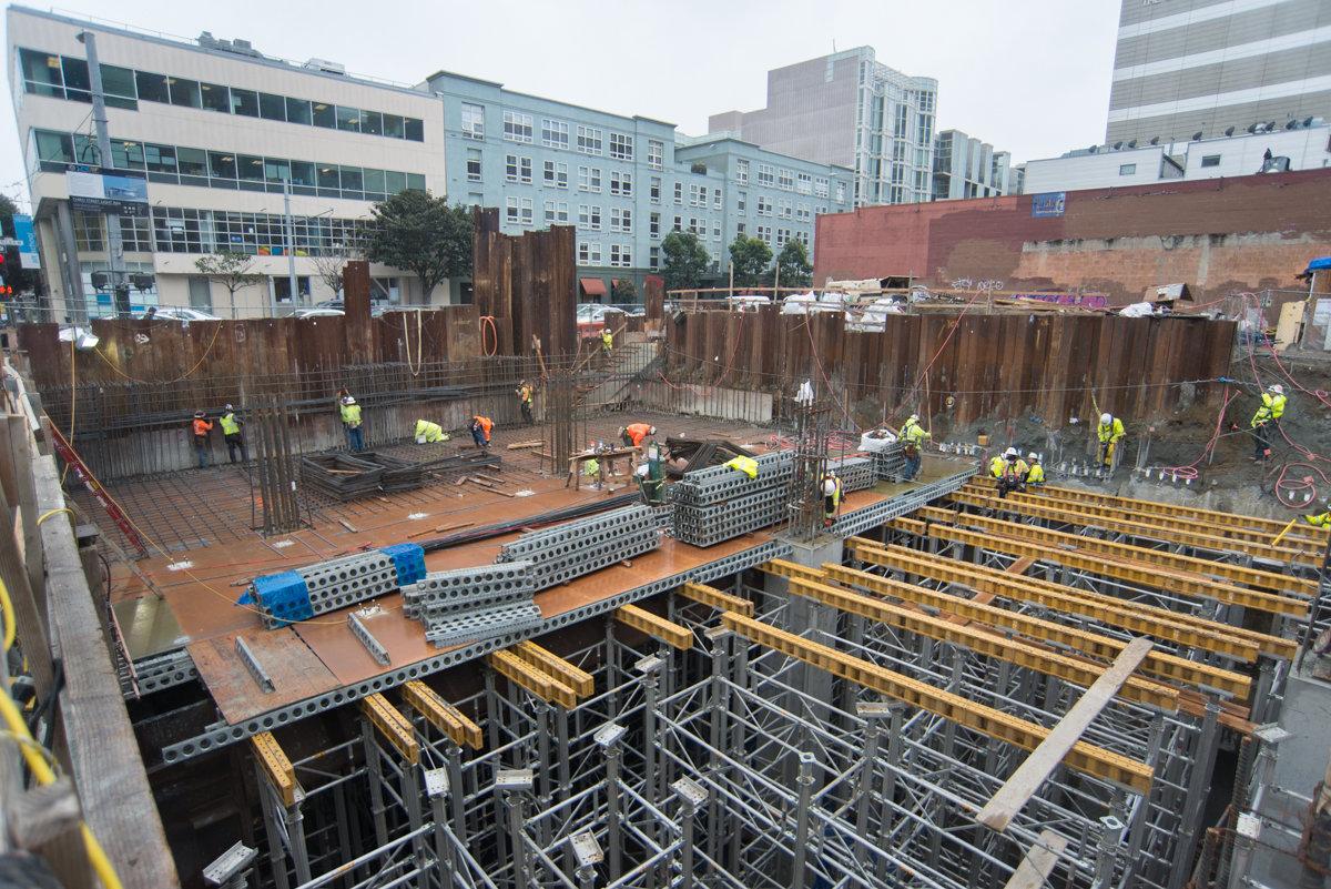 Construction workers and equipment inside underground station excavation with steel reinforcement and wooden shoring.