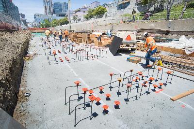 Construction workers installing rebar on concrete foundation in excavated trench for transit project.