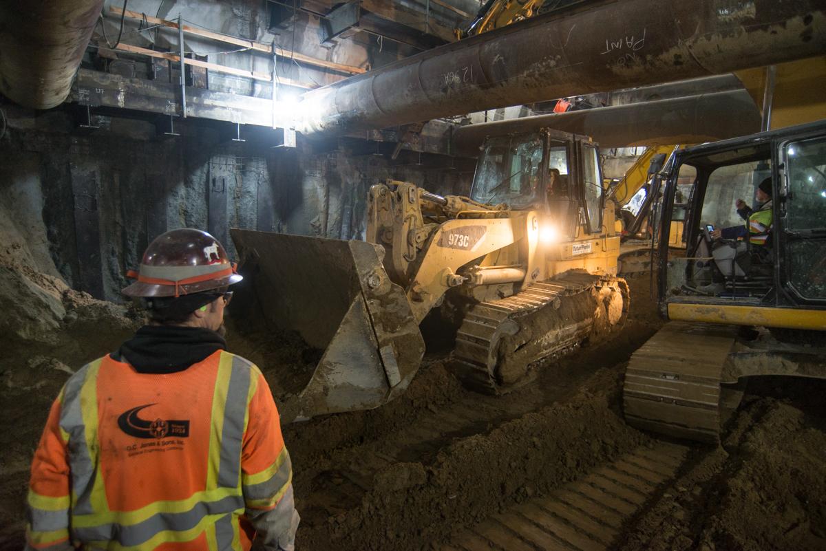 Worker in safety gear observing bulldozer and excavator operating in underground station construction site.