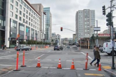 Urban street with traffic cones blocking lanes, construction barriers, and city buildings in background.