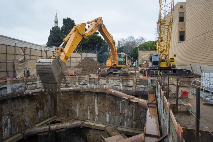 Yellow excavator operates at construction site with tunnel boring machine retrieval shaft and wooden support structures.