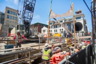 Construction workers in safety gear at underground transit station excavation site with steel framework and city buildings visible above ground.