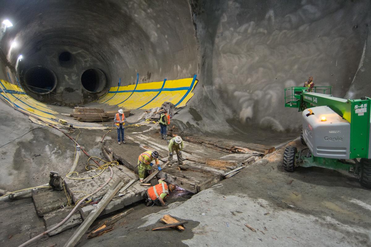 Workers in safety gear clear debris from tunnel floor near large concrete pipes and yellow cables.